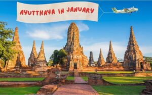 A scenic view of the ancient brick ruins of Wat Chaiwatthanaram in Ayutthaya under a bright blue sky, featuring a digital overlay of a small plane pulling a banner that reads "AYUTTHAYA IN JANUARY.