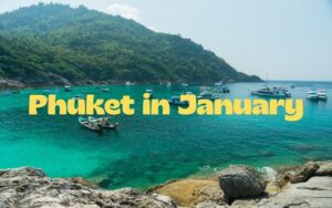 A scenic tropical bay in Phuket featuring turquoise waters filled with anchored boats and a lush green hillside background, with the text "Phuket in January" overlaid in yellow.