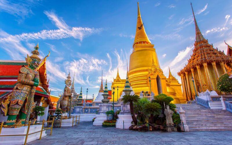 A stunning view of Wat Phra Kaew in Bangkok, featuring the golden stupa and traditional Thai temple structures under a vibrant blue sky with scattered clouds