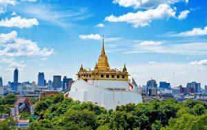 Wat Saket with its golden chedi rising above lush greenery and Bangkok’s skyline under a bright blue sky.